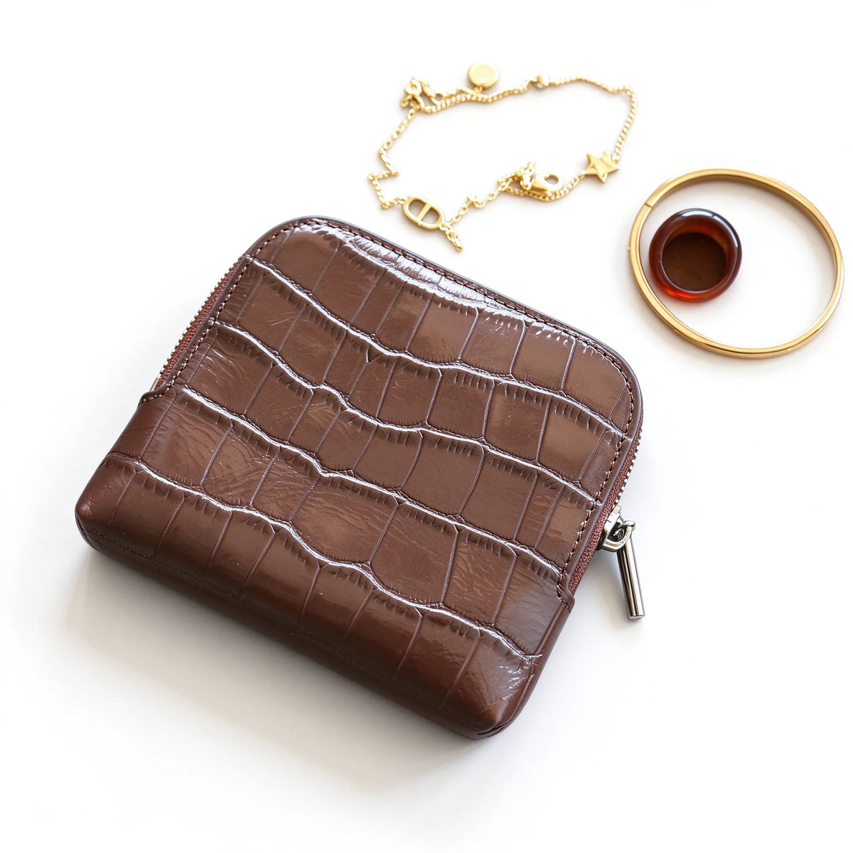 Brown crocodile-textured leather jewelry pouch with a zip-around opening, shown beside bracelet and ring props.