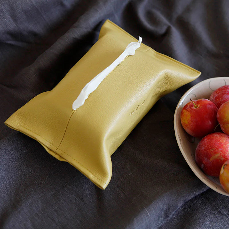 Yellow leather tissue holder with a central opening on dark fabric beside a bowl of apples