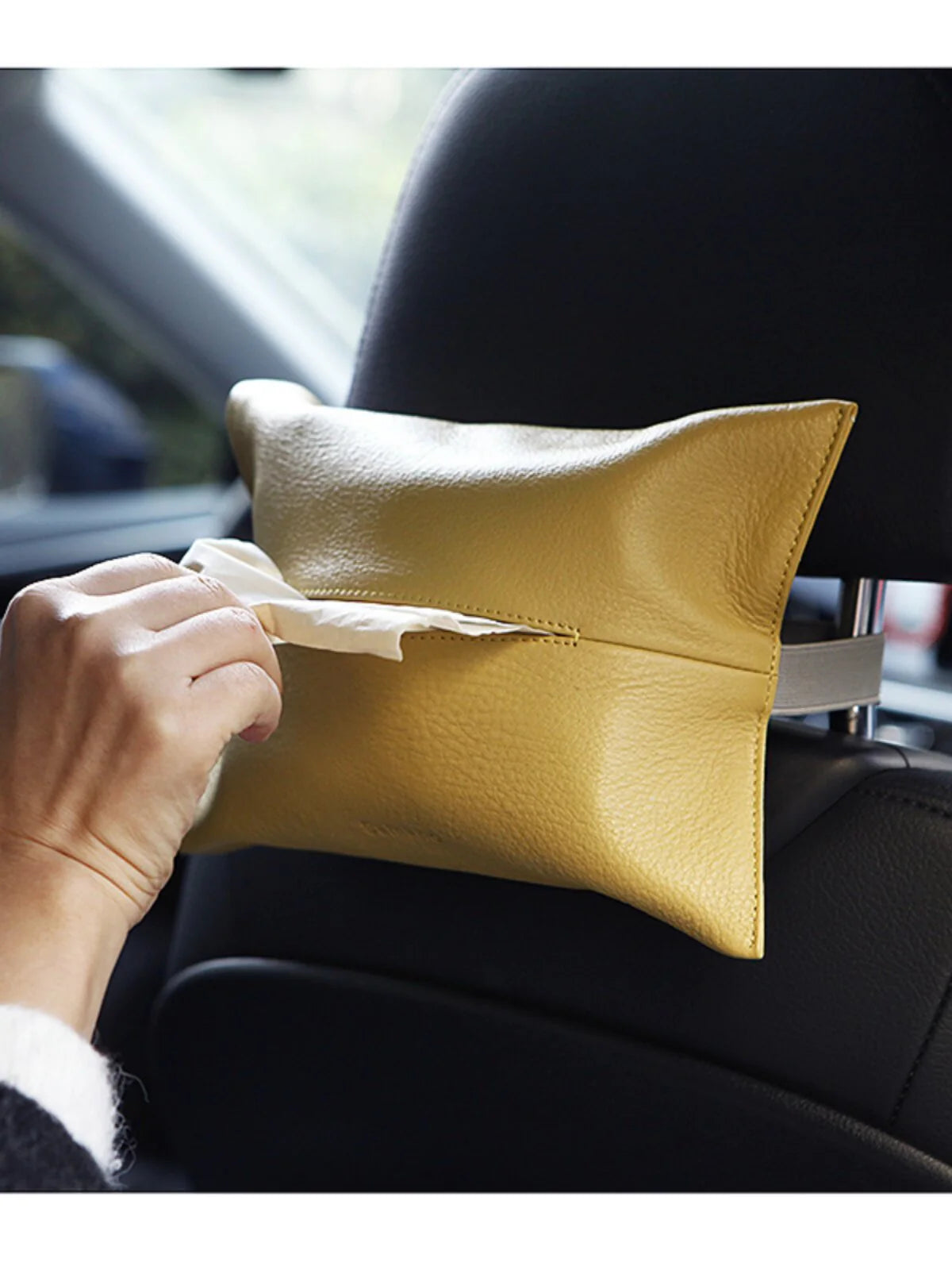 Yellow leather car tissue holder secured behind a headrest while a hand pulls a tissue
