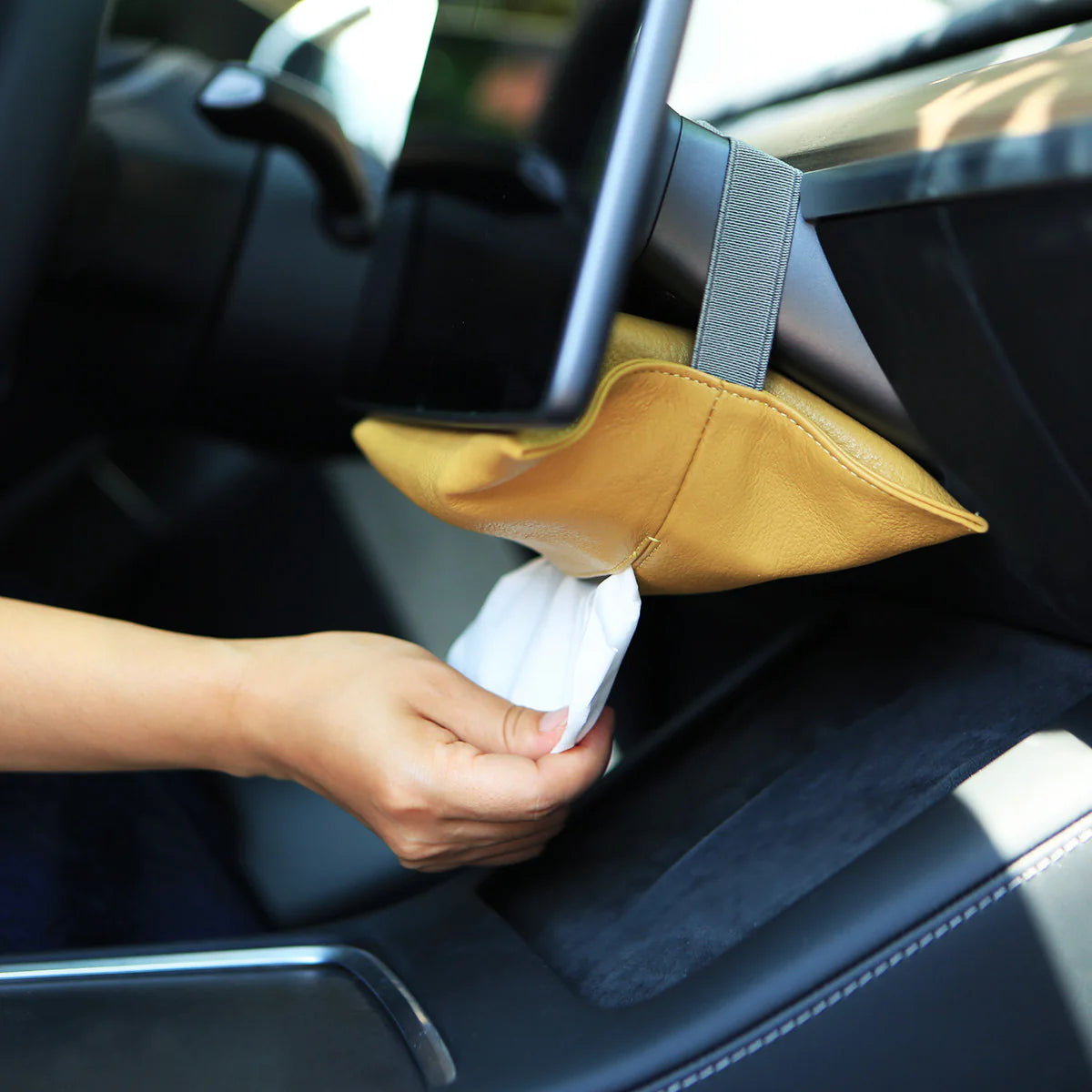 A hand pulling a tissue from a yellow leather holder beneath an in-car display