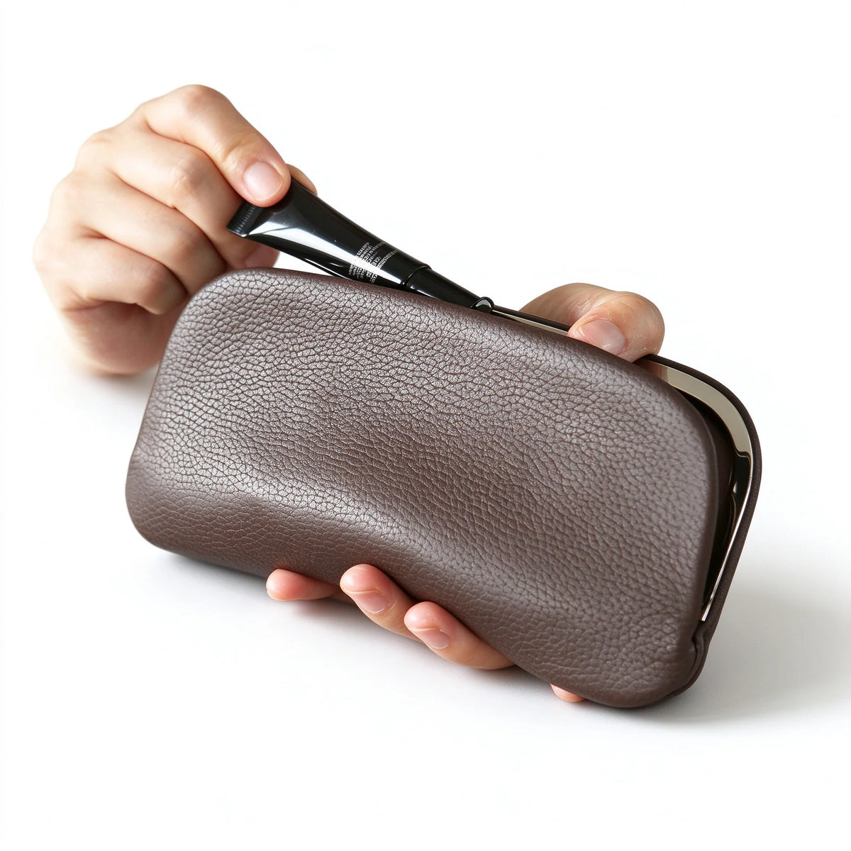 Dark brown leather clutch pouch held open while a cosmetic tube is placed inside
