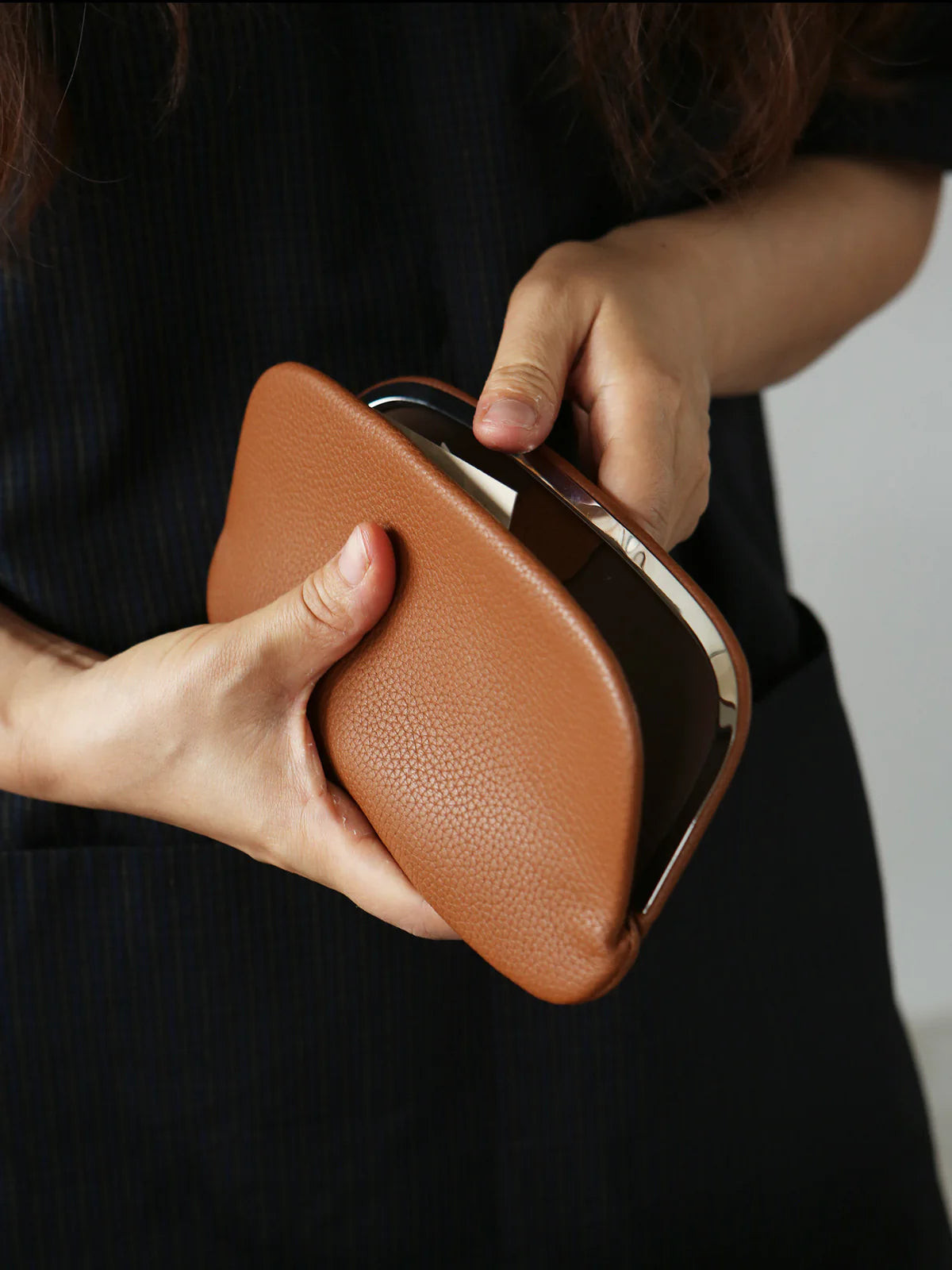 A person holding an open tan leather metal-frame clutch pouch against a black outfit