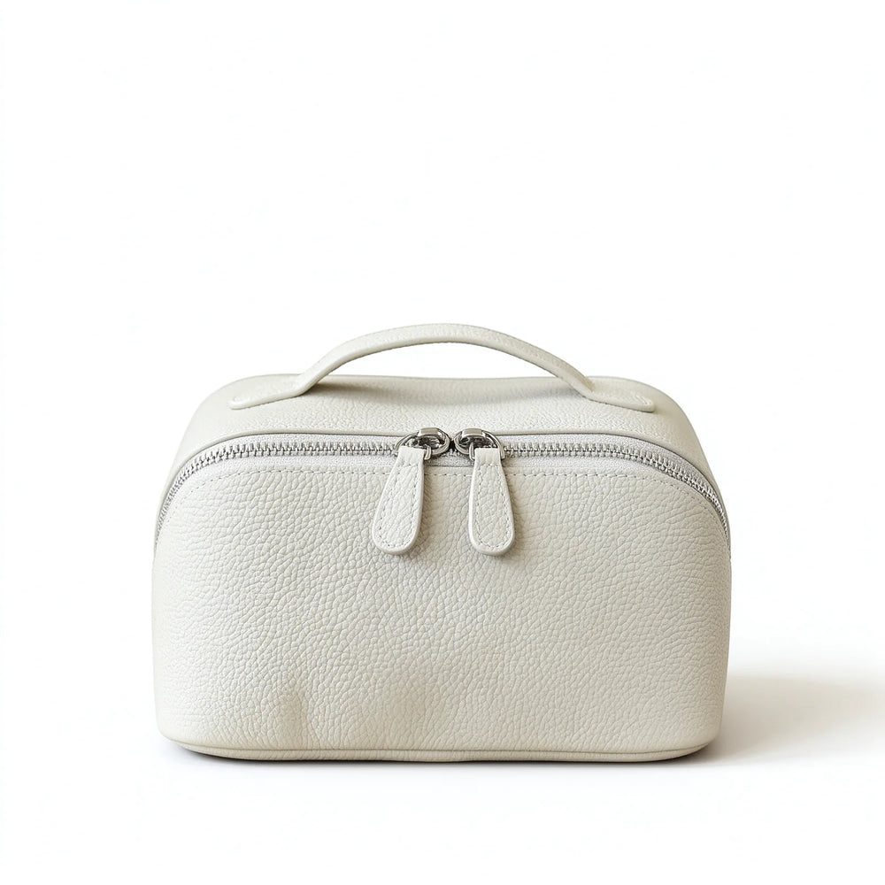 Front view of an off-white leather travel makeup bag standing upright with a top handle and double zip pulls.