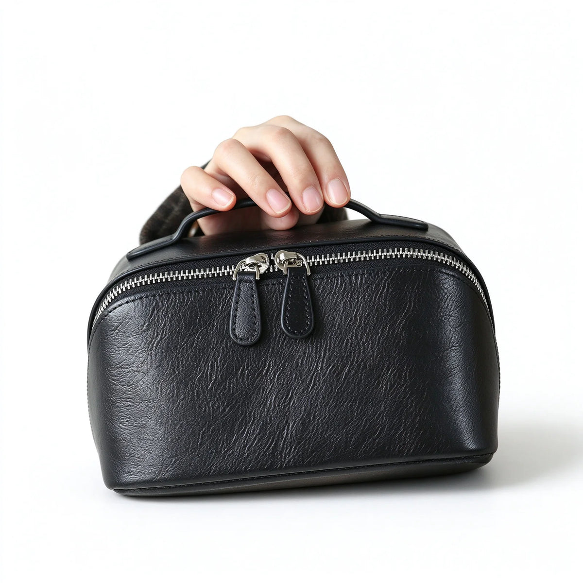 Black leather travel makeup bag held by its top handle, with double leather zip pulls and a pebbled front panel.