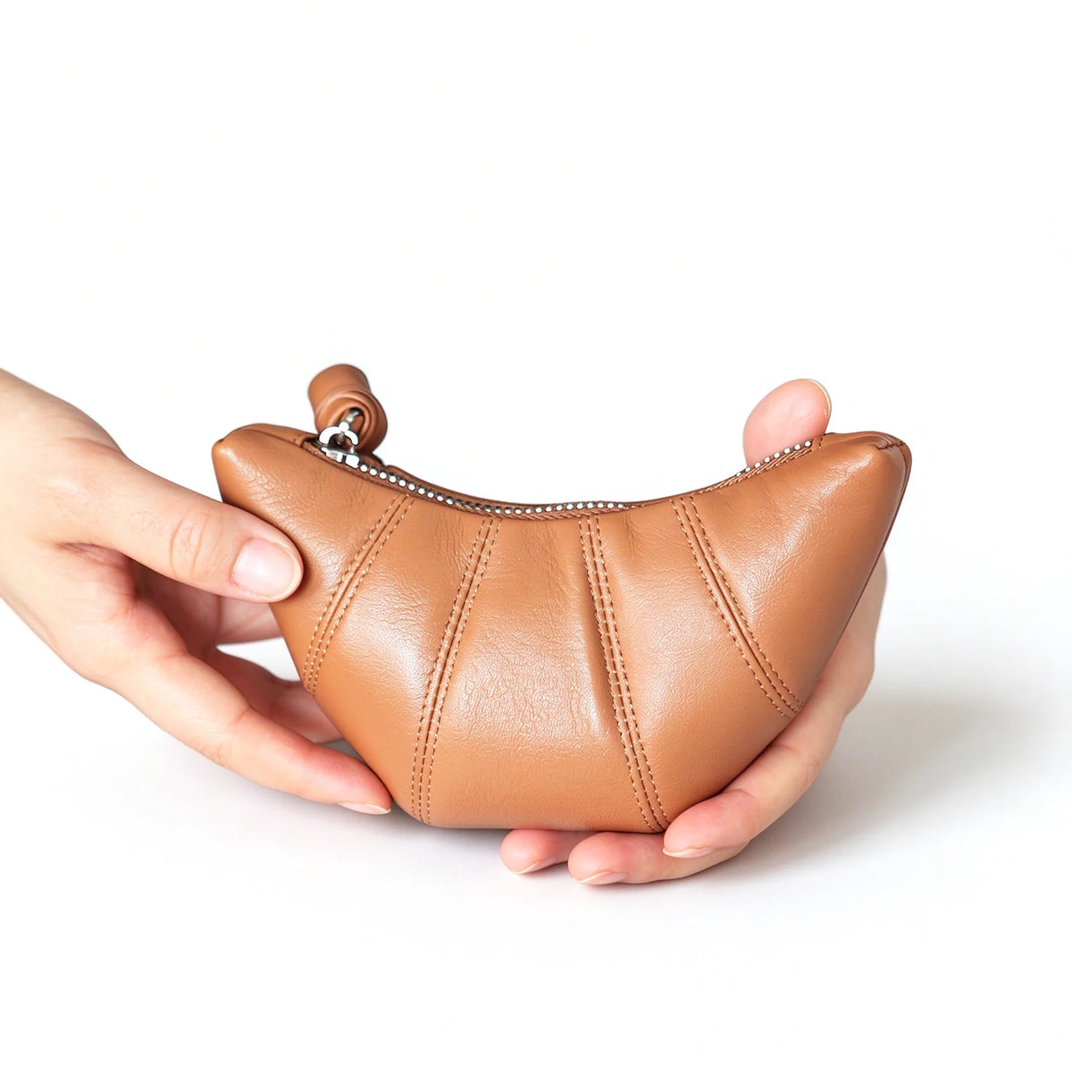 Tan crescent-shaped leather makeup pouch held in both hands, showing stitched panels and a curved zipper.